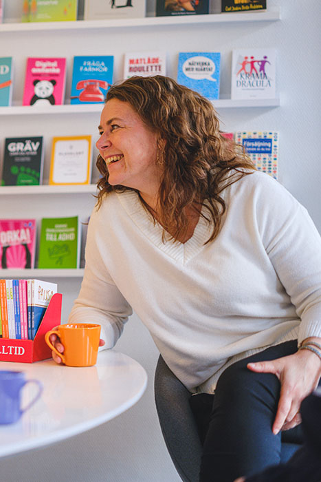 Författaren och yogainstruktören Ulrika Borén sitter på förlaget med en kopp kaffe i handen och pratar med förläggaren Helene. Bakom Ulrika syns en vägg med kunskap på nolltid-böcker. Foto av Crelle