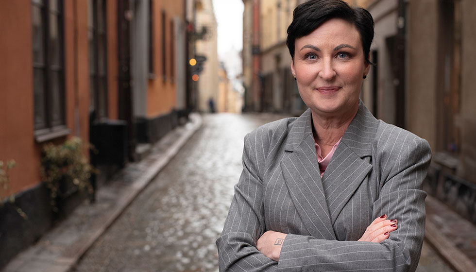 Författaren och psykologen Sandra Lindström. På bilden står hon i en kritstrecksrandig, grå kavaj med armarna i kors. I bakgrunden syns Gamla stan i Stockholm